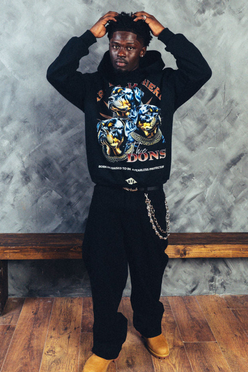 Person wearing a black hoodie with colorful graphics, standing against a textured gray wall.