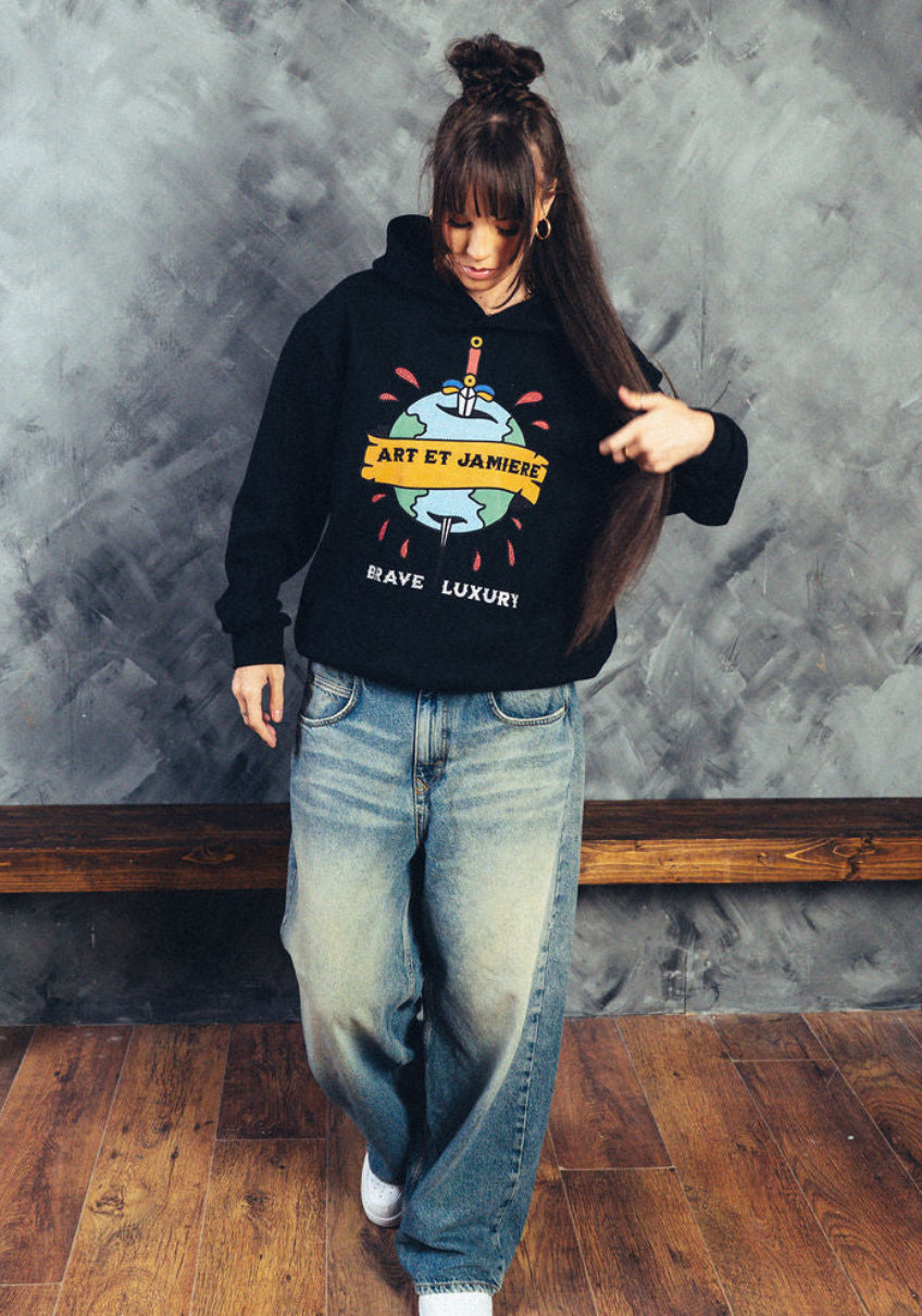 Person wearing a black hoodie with colorful graphics and text, standing against a textured gray wall with a wooden bench.