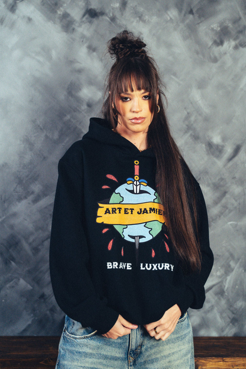 Person wearing a black hoodie with a graphic design against a textured gray wall.